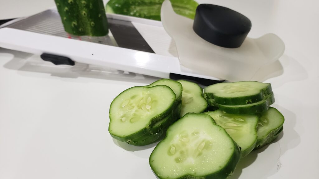 Pickling cucumbers are cut in slices and laying on a counter in front of a mandoline