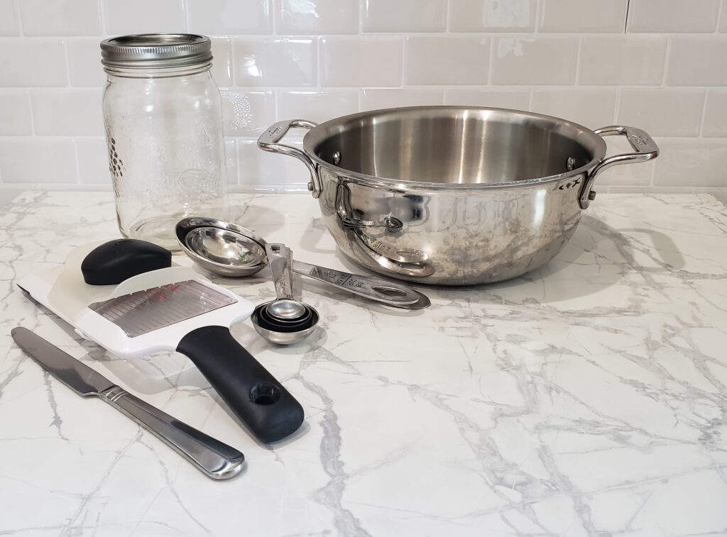 pickling tools sit on a white counter top including a stainless steel pot, a 1L mason jar and some measuring spoons