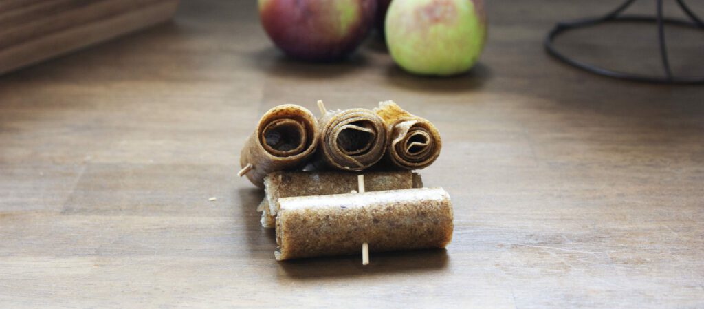 apples sit in the background while apple fruit leather or apple fruit roll ups are rolled in the foreground of the photo