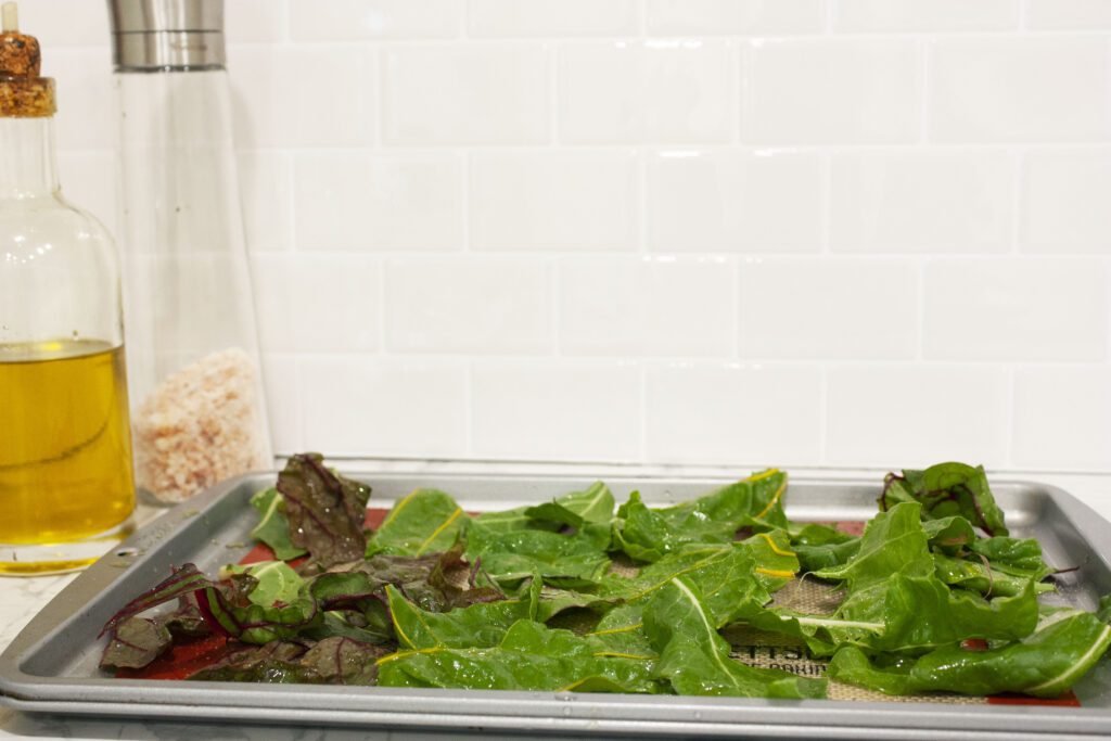 a baking sheet is filled with cut up swiss chard leaves which are covered in salt and oil
