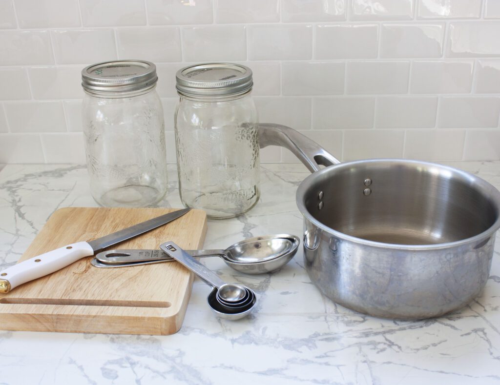 mason jars, measuring spoons, a pot, knife and cutting board sit on a white counter as equipment needed to make pickled beans
