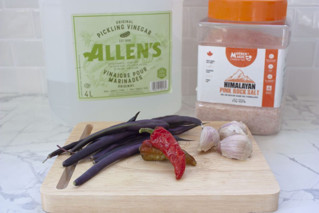 white vinegar, pink salt, purple beans, garlic cloves and red hot peppers sit on a wooden cutting board