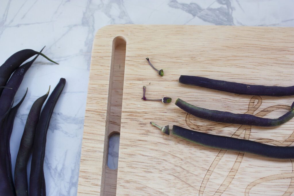 3 purple beans sit on a wood cutting board with their stem cut off