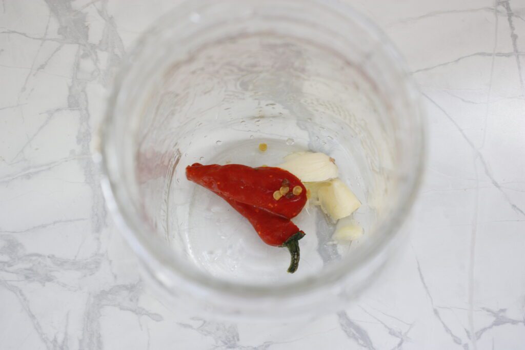 a glass mason jar sits open on a white counter with a crushed garlic clove and a red pepper at the bottom for pickling beans