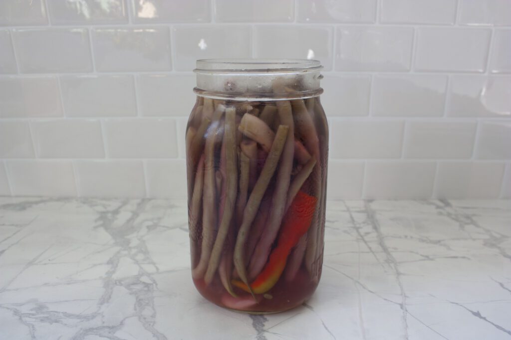 a glass jar filled with pickled beans and a hot pepper sit in brine on a white counter
