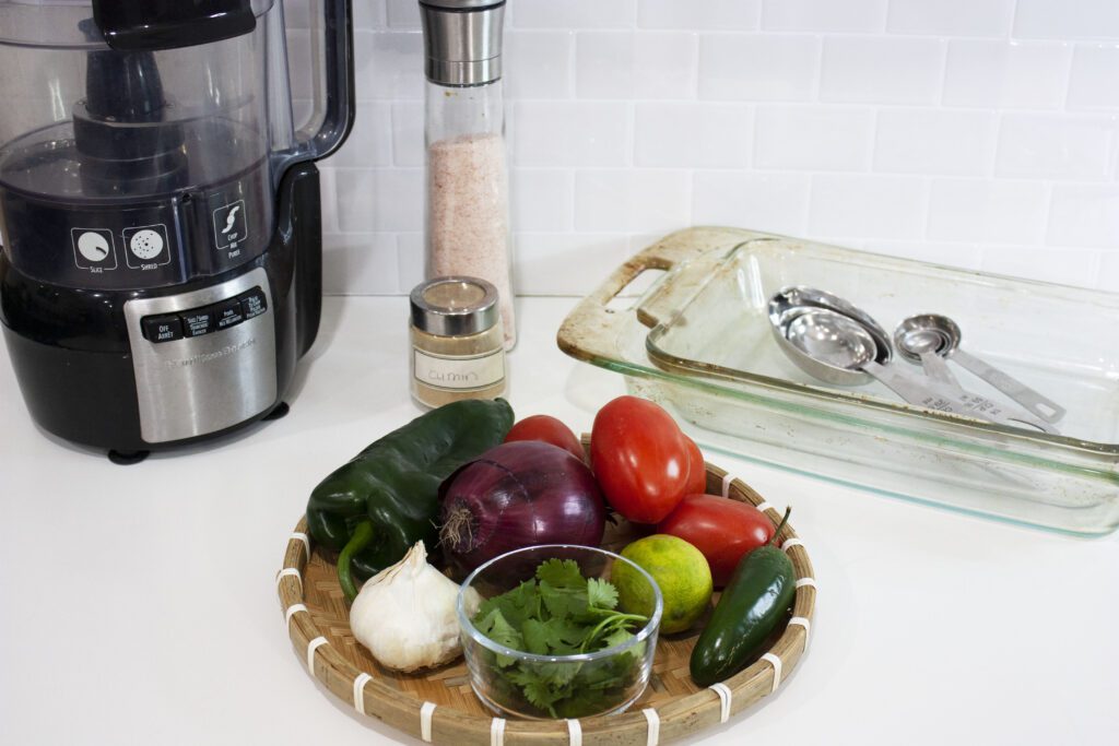 the fresh ingredients of salsa like tomatoes, onion and peppers sit on a counter with a food processor and other equipment