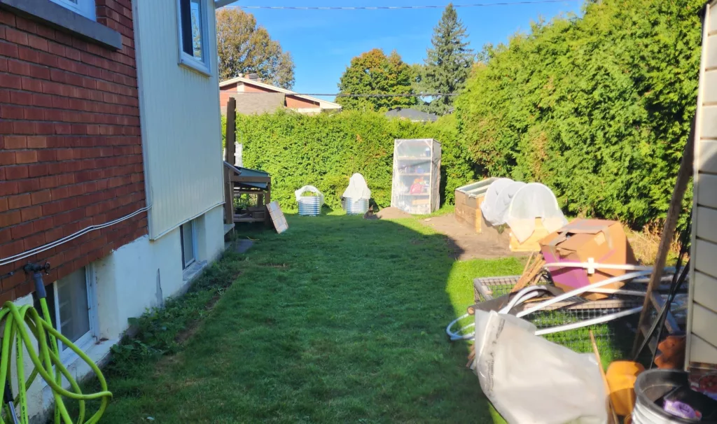 a sideyard of a house is shown with shade close to the house and a bunch of gardens and junk on the sides