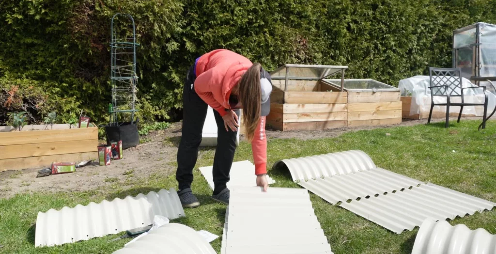 a woman in a red sweatshirt and black pants is building a white metal raised bed garden with panels laid out