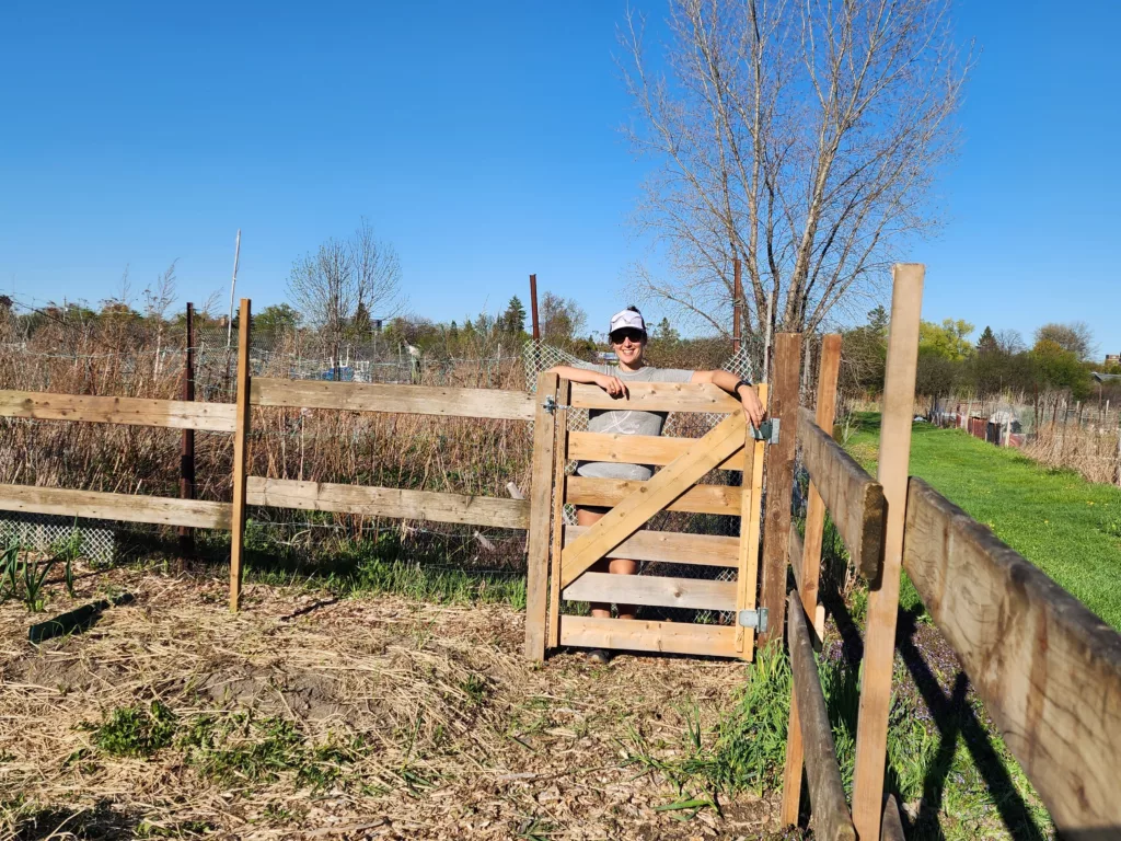 a white woman poses behind a wooden gate with a wood fence surrounding an allotment. A blue sky and leafless tree is in the background