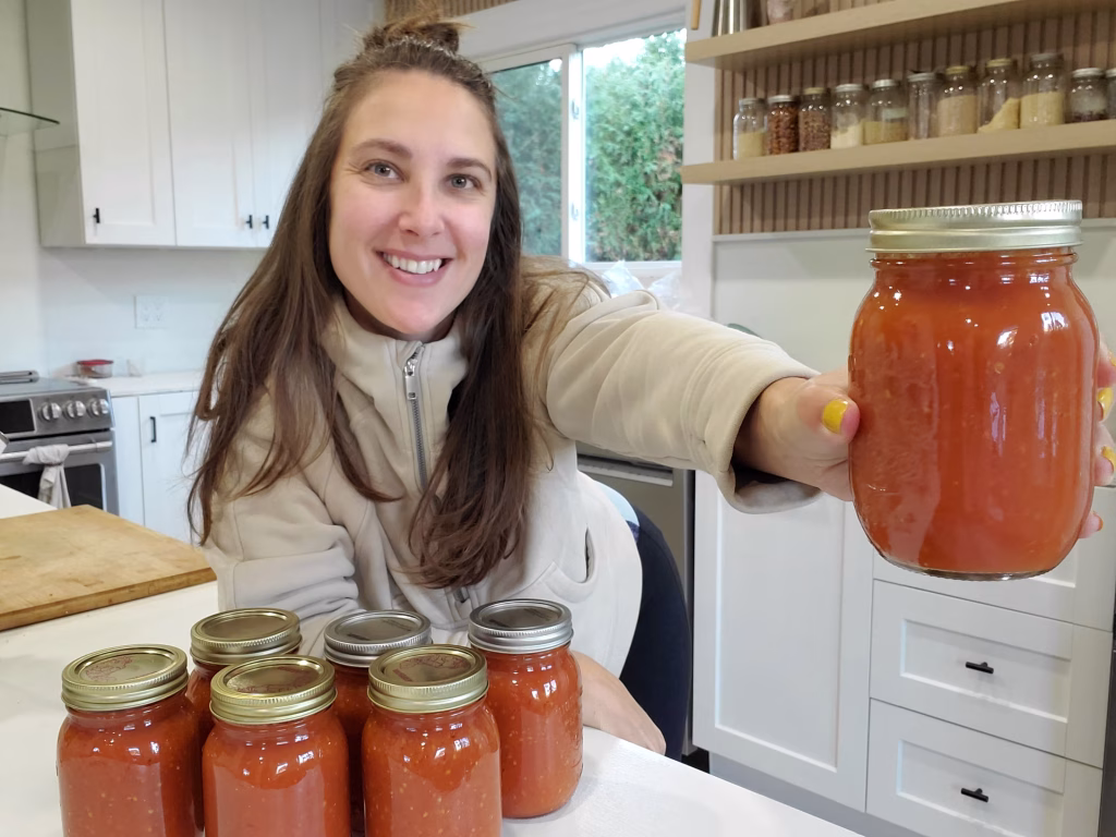 a woman holds a jar of tomato sauce with 6 jars infront of her that she made from water bath canning